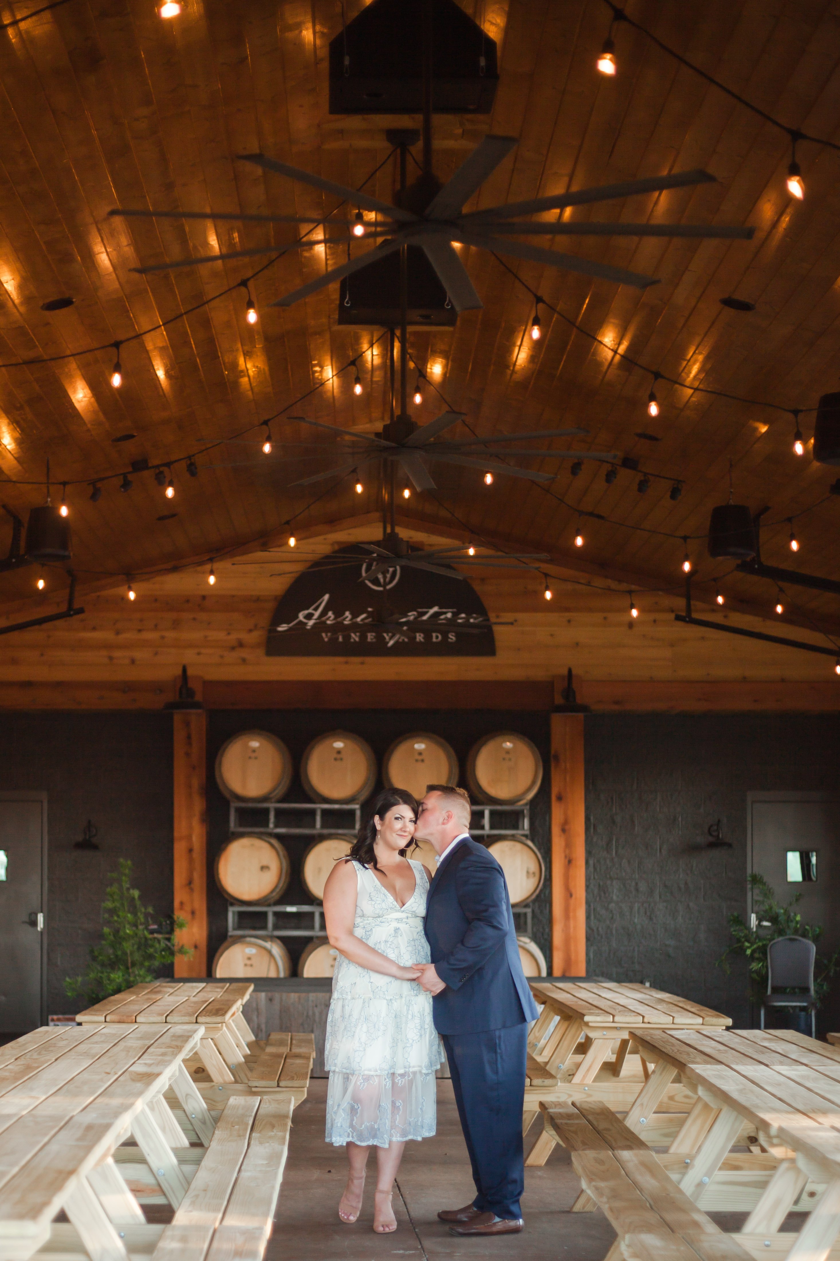 guy in navy suit kissing girl in white dress in front of wine barrels